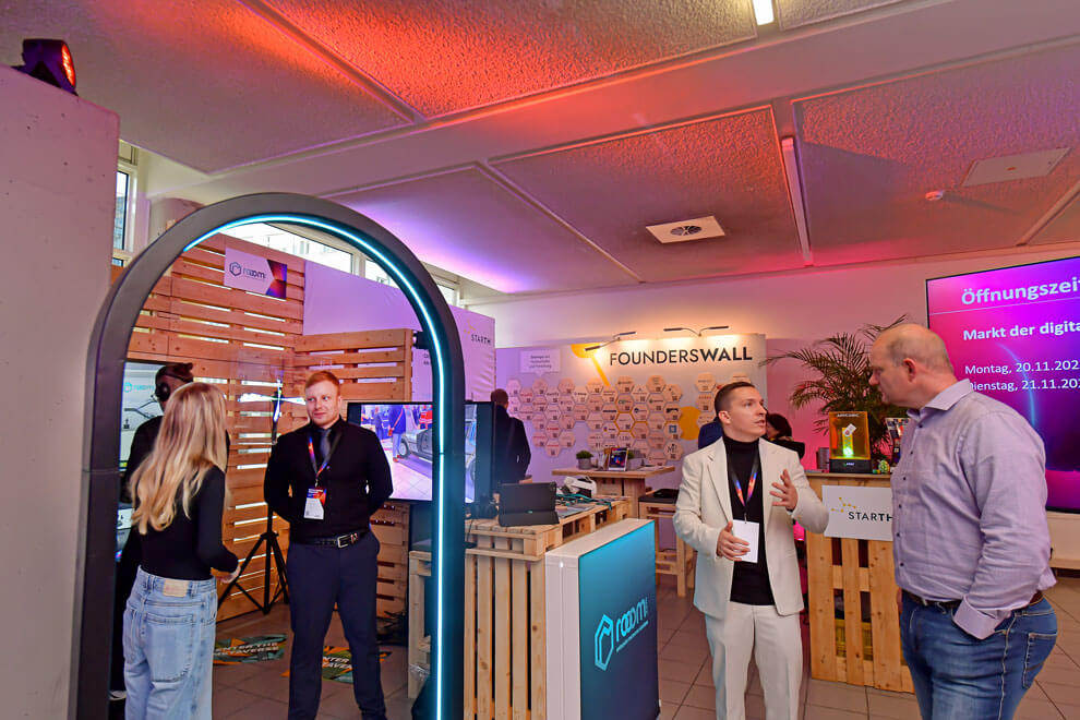 Menschen im Gespräch an einem modernen Messestand mit Holz- und Lichtelementen. Im Hintergrund sind eine Founderswall, Displays und Informationen zu einem Event zu sehen