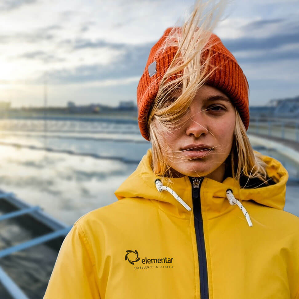 Porträt einer Frau mit orangefarbener Mütze und gelber Jacke vor einer Wasseranlage