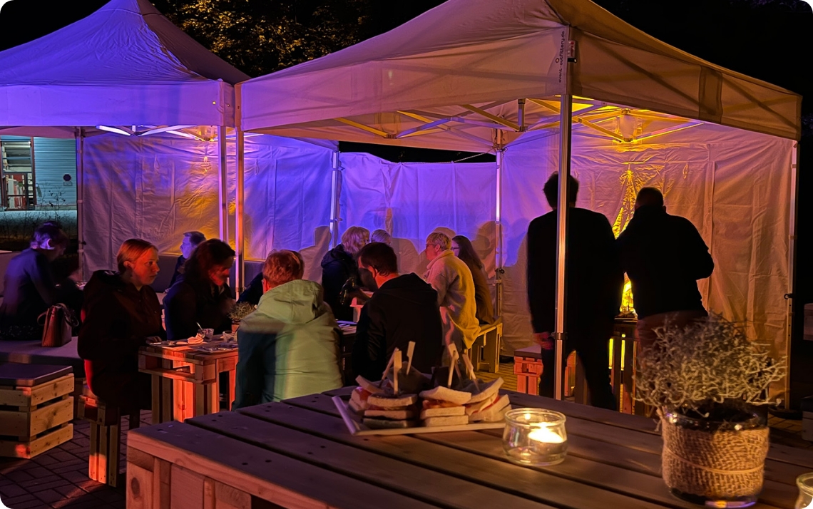 Abendveranstaltung in Pavillions mit stimmungsvoller Beleuchtung. Warme Lichtquellen und farbige Akzente schaffen eine einladende Atmosphäre. Lockeres Setting mit Stehtischen und Sitzbereichen fördert Austausch und Gespräche