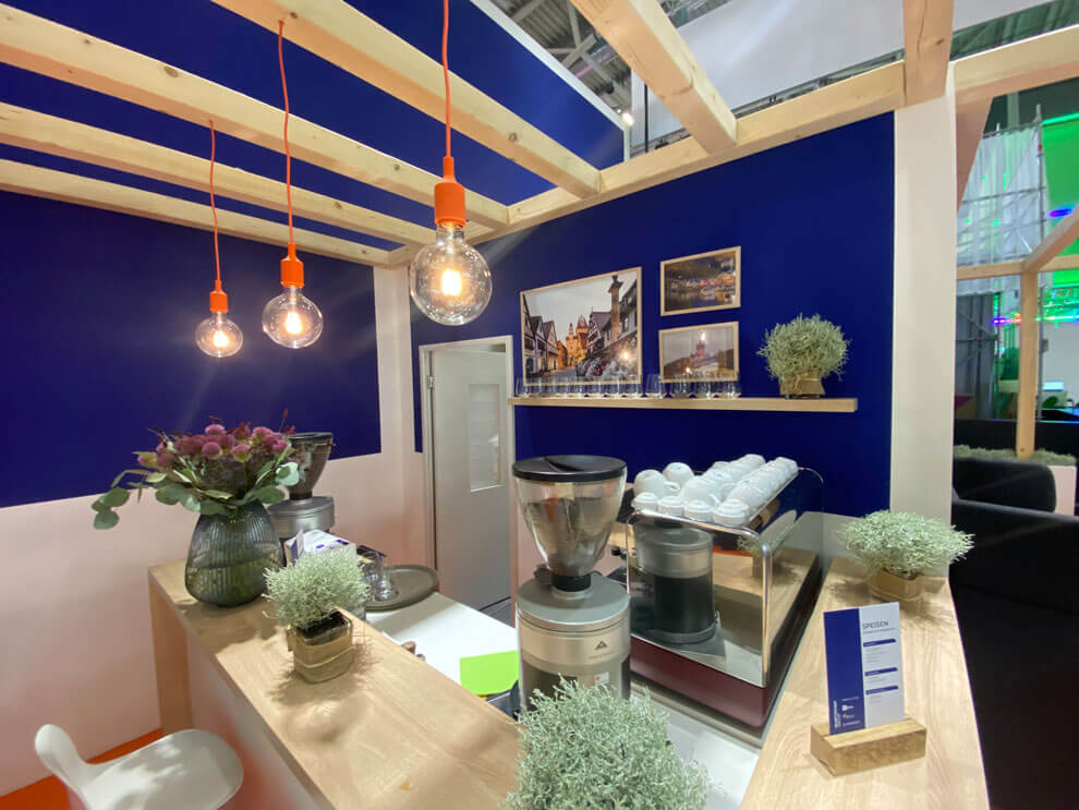 Kompakter Barbereich im Messestand. Warme Holzoberflächen, blaue Wandflächen und dekorative Beleuchtung schaffen eine einladende Atmosphäre