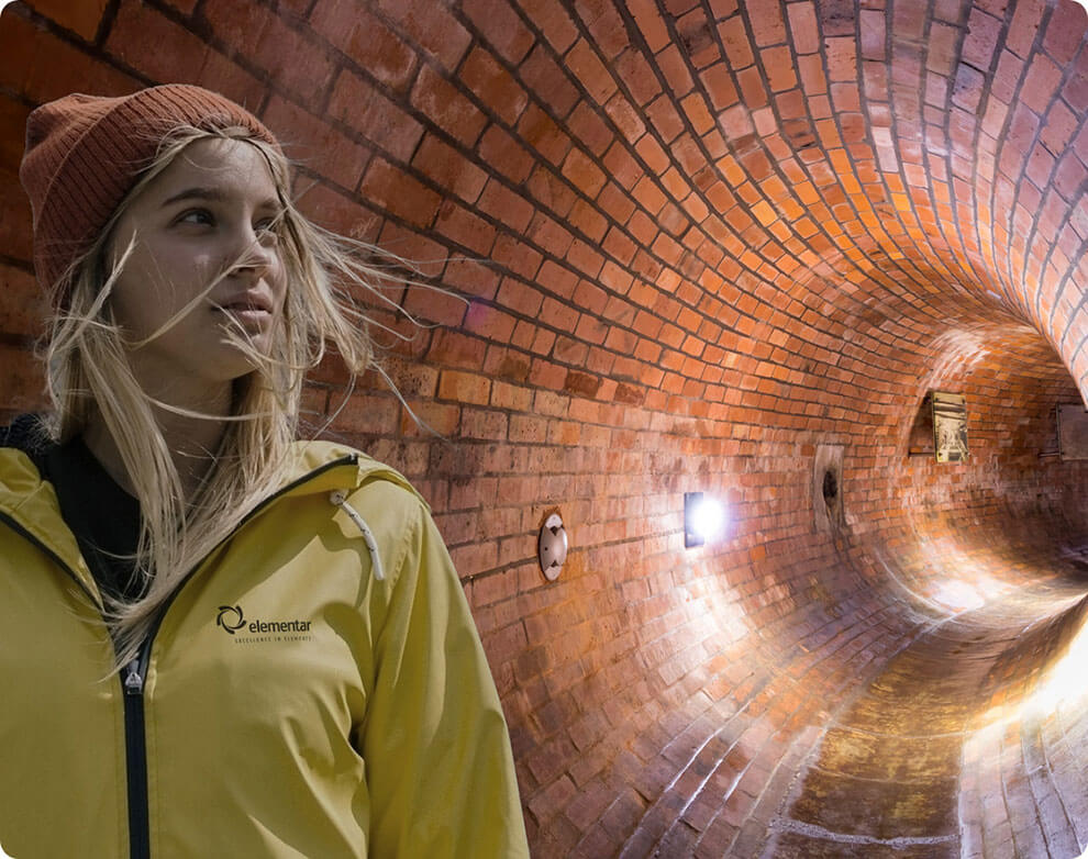 im Vordergrund ist eine Frau mit gelber Elementar-Regenjacke und orangener Mütze. Im Hintergrund ist ein beleuchteter Abwasserkanal