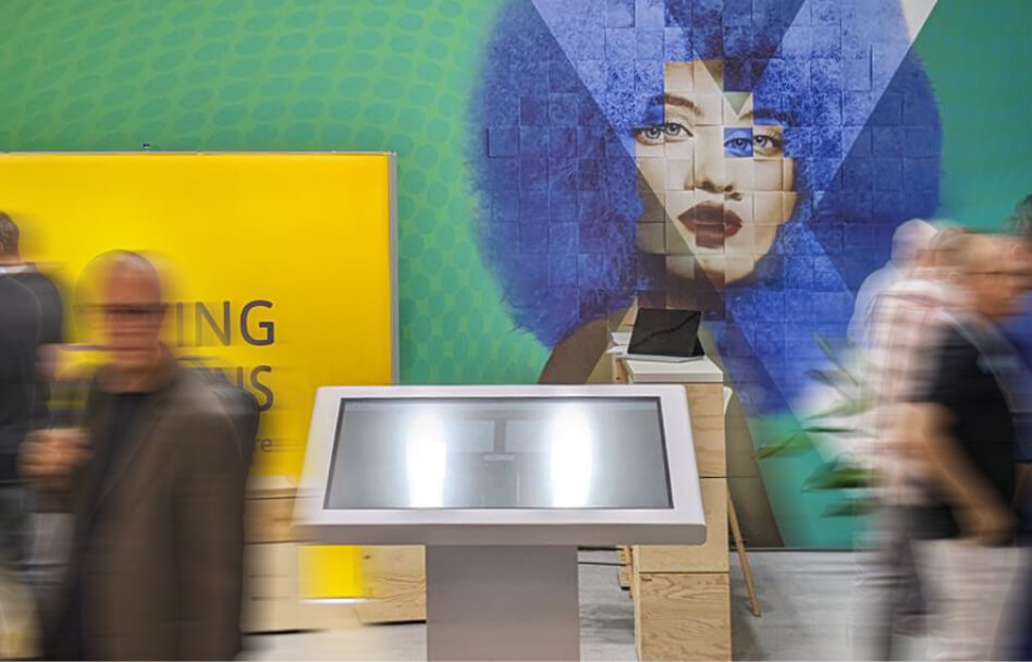 Besucher bewegen sich vor einem interaktiven Display auf einem Messestand mit großformatiger Wandgrafik im Hintergrund
