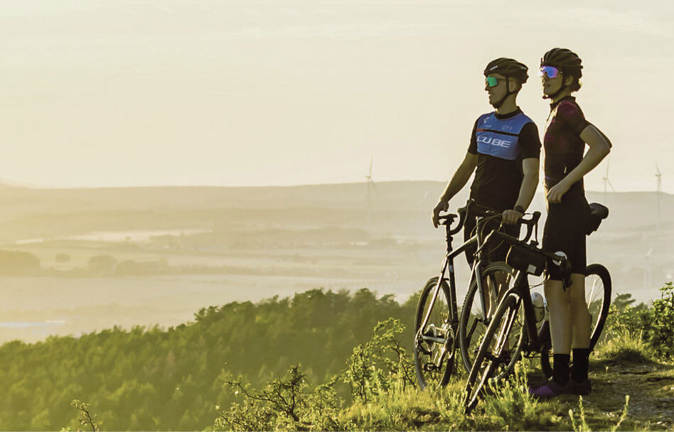 Radfahrer mit Rennrädern auf Aussichtspunkt in Thüringens Süden mit Blick über Landschaft und Windräder bei Sonnenuntergang