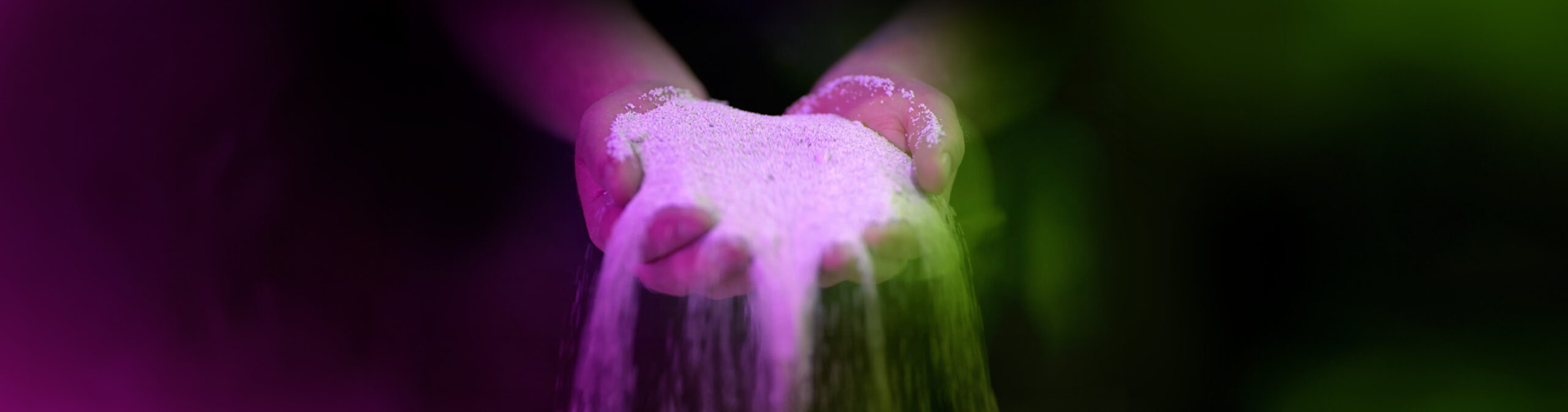 AKT_KOM_SchubSa-IIJ_Webheader_fl_01 Zwei Hände halten feinkörniges, helles Material, das langsam zwischen den Fingern hindurchrieselt, beleuchtet von einem farbigen Lichtverlauf in Magenta und Grün