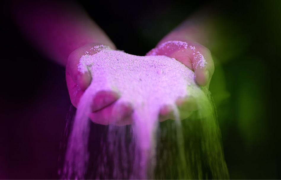 AKT_KOM_SchubSa-IIJ_Webheader_fl_03 Zwei Hände halten feinkörniges, helles Material, das langsam zwischen den Fingern hindurchrieselt, beleuchtet von einem farbigen Lichtverlauf in Magenta und Grün