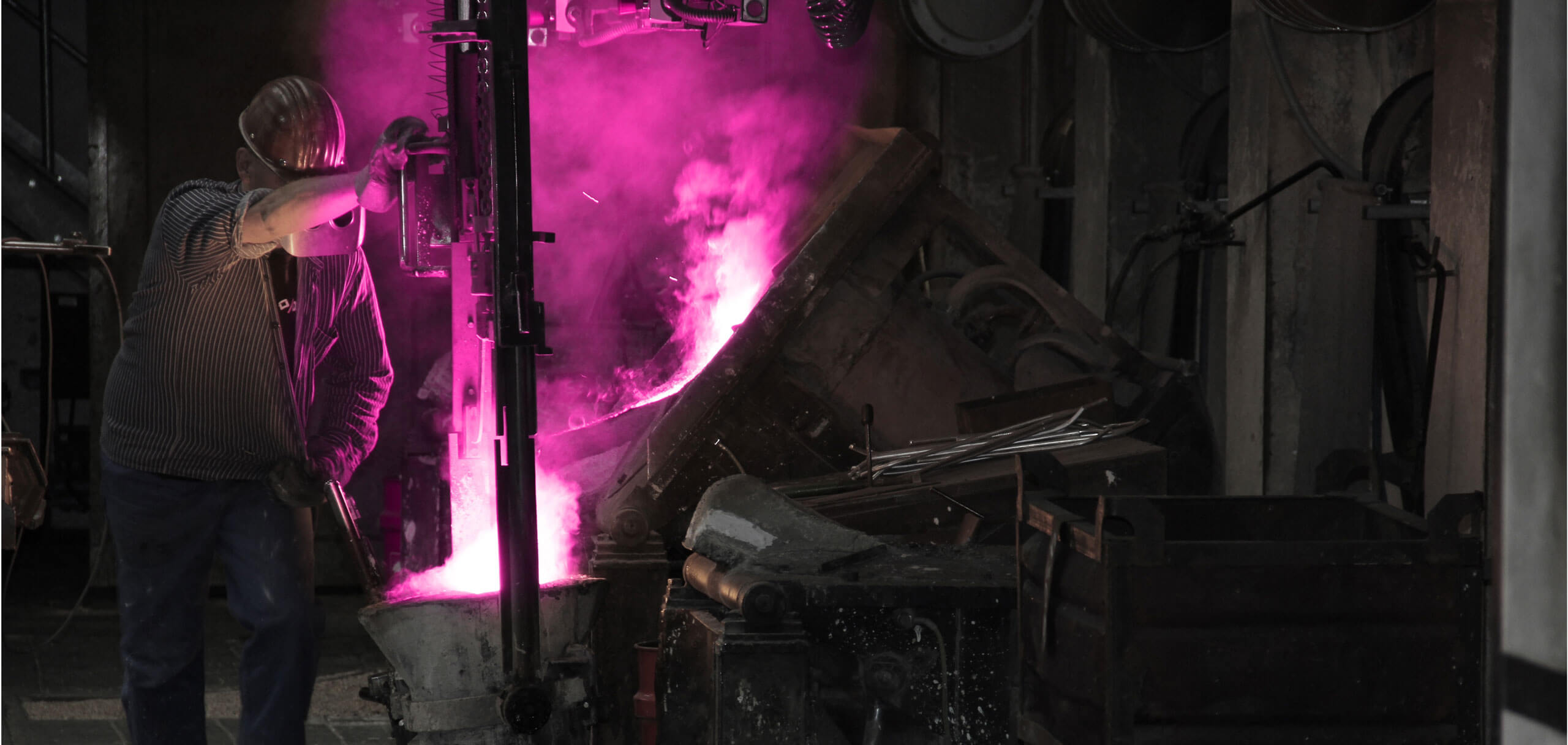Ein Arbeiter in Schutzkleidung bedient eine Gießanlage in einer Industriehalle, während glühendes Metall mit intensiv magentafarbenem Licht und Rauch in eine Form gegossen wird