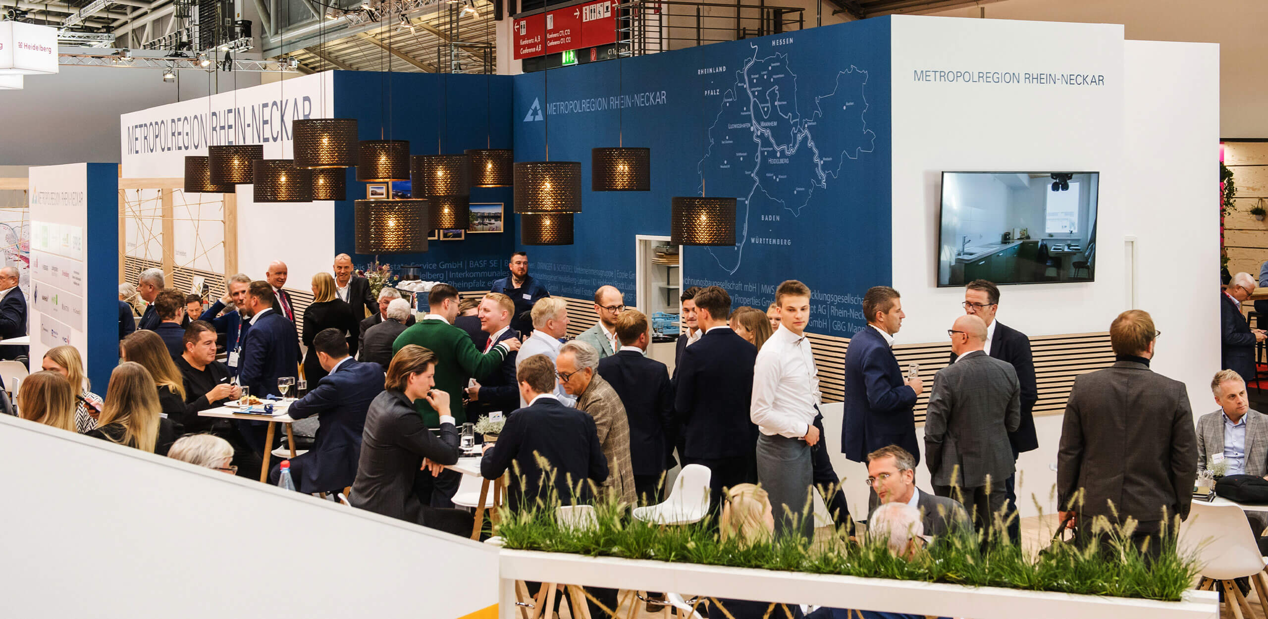 Besucherinnen und Besucher stehen und sitzen an Tischen auf dem Messestand der Metropolregion Rhein-Neckar und führen Gespräche