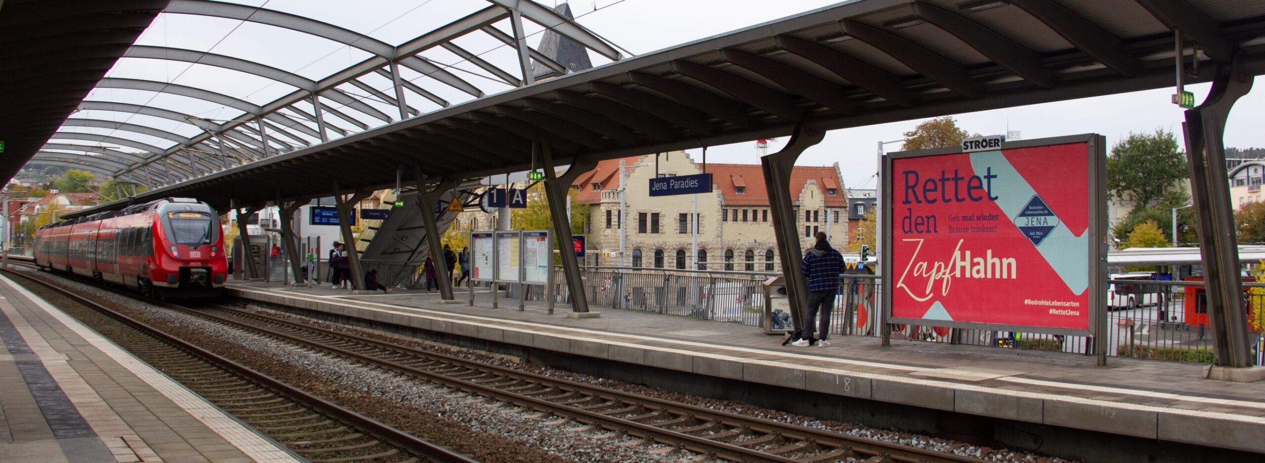 Ein Bahnsteig in Jena mit einfahrendem Regionalzug. Auf dem gegenüberliegenden Gleis hängt ein großes Plakat der Kampagne „Rettet den Zapfhahn“
