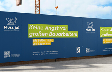 Blauer Bauzaun mit Kampagnenmotiv „Muss ja!“ und der Botschaft „Keine Angst vor großen Bauarbeiten! Die beißen nicht, die bauen nur.“ zur Information und Beruhigung von Bürgerinnen und Bürgern an einer Baustelle