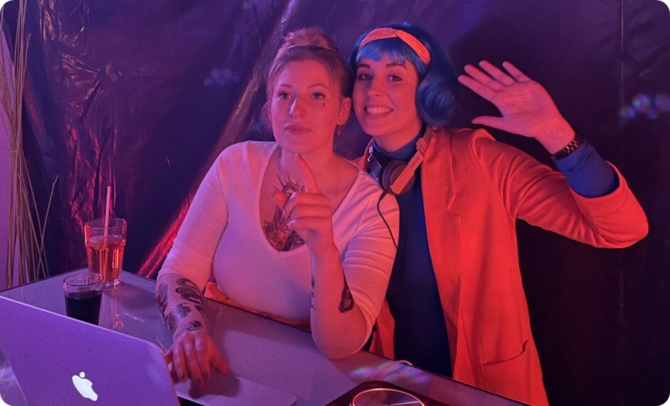 12 Zwei Mitarbeiterinnen sitzen an einem Laptop in farbig beleuchteter Eventumgebung, eine winkt in die Kamera, als Moment für Kreativität, Teamspirit und gemeinsame Aktionen