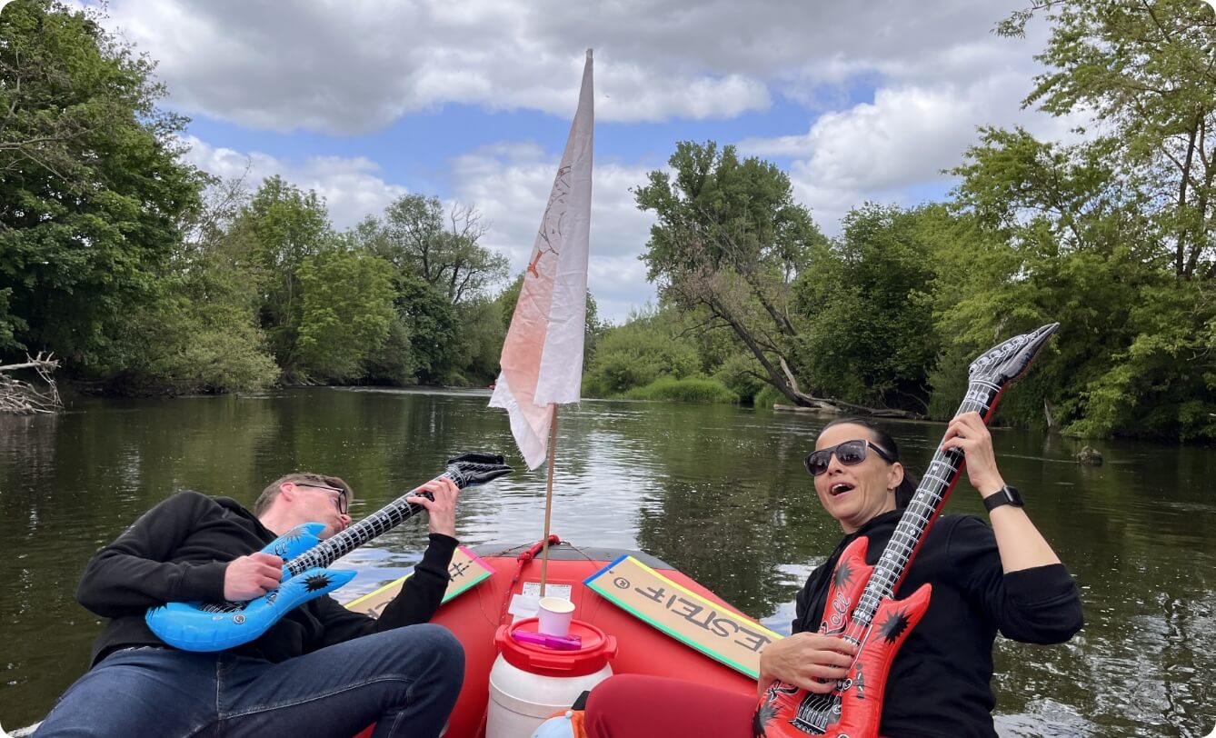 Zwei Mitarbeitende sitzen in einem Schlauchboot auf einem Fluss und spielen scherzhaft Luftgitarre, als humorvolles Bildmotiv für Teamgeist, Spaß und gemeinsame Erlebnisse