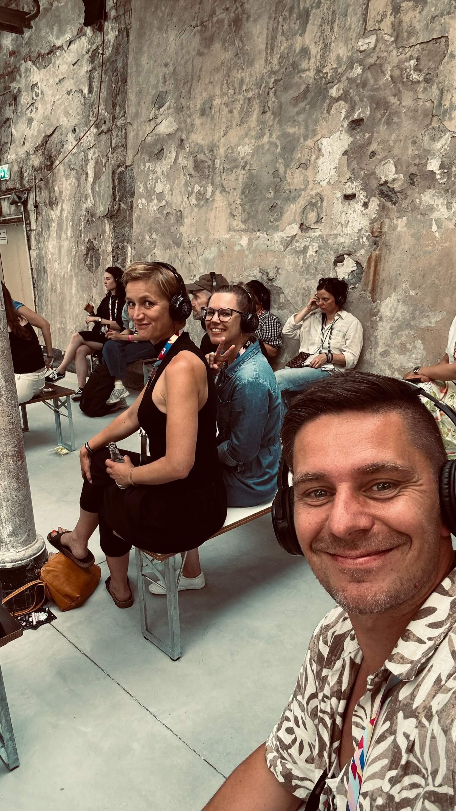 Mehrere Mitarbeitende sitzen mit Kopfhörern bei einer Veranstaltung in industrieller Umgebung, ein Teilnehmer macht ein Selfie, als Bildmotiv für Austausch, Inspiration und gemeinsames Lernen